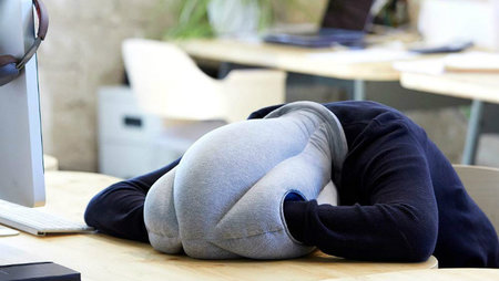 A person wearing an OSTRICH PILLOW travel pillow and sleeping at their desk